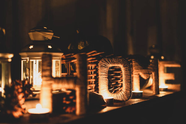 Decorative composition with inscription HOME, pine cones, candles and lanterns. Letters covered with twine in candlelight. Selective focus on letter O.