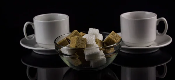 Two white coffee cups with white and brown sugar cubes on a dark background