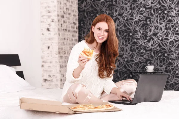 Kızıl saçlı kız yatakta dizüstü bilgisayarda çalışan ve pizza yemek
