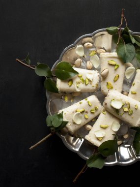Pista kulfi, Hint dondurma plaka hizmet buz küpleri, Antep fıstığı, gül yaprakları, siyah arka plan, üstten görünüm üzerinde yeşil yaprakları ile ağaç dalları üzerinde. Geleneksel Rajasthani Hint Mutfak.