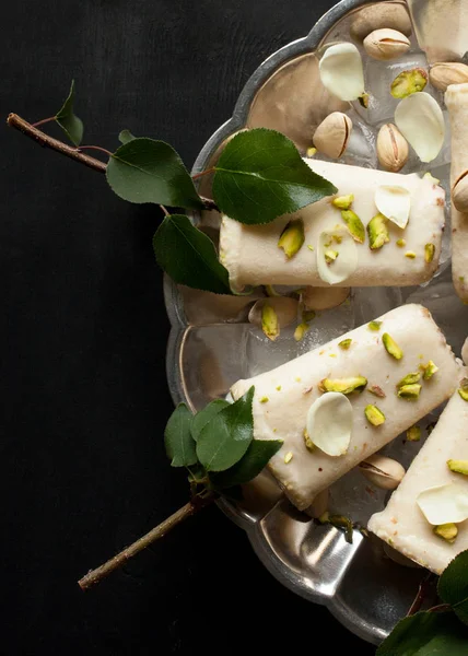 Pista kulfi, Hint dondurma plaka hizmet buz küpleri, Antep fıstığı, gül yaprakları, siyah arka plan, üstten görünüm üzerinde yeşil yaprakları ile ağaç dalları üzerinde. Geleneksel Rajasthani Hint Mutfak.
