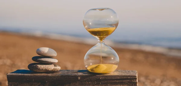 Sandglass on a wooden board with three stones in the form of a pyramid, a modern hourglass on a beach with golden sand against the background of the sea