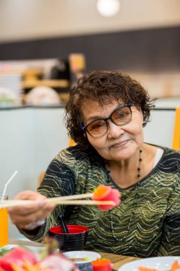 Asya 'lı yaşlı kadın turist elinde çatalla gülümseyerek yerel yemekleri veya Japon yemeklerini gösteriyor..