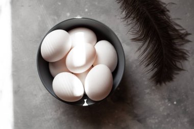 Tabak yumurtayla dolu. Tüyler yakınlarda. Beton zeminde dörtten fazla yumurta var. Fotoğraf, gri tabak ve tavuk yumurtası..