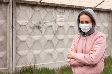 Kapüşonlu ve beyaz maskeli bir kız. Pembe ceketli, arka planda beton bir çit, çimenler ve ağaçlar. Karantina zamanı. Portre fotoğrafı, yazı için fotoğraf..