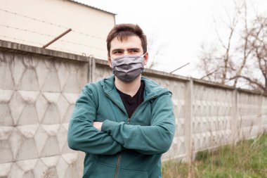 Açık havada gri tıbbi maskeli bir adam. Elleri bağlı. O, yeşil bir kazak giydi. Arka planda gri beton bir çit, çimenler ve ağaçlar var. Karantina zamanı.