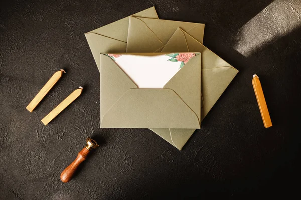 Blank paper envelopes with golden wax seal, stamp, spoon and postcard on black paper background. Mockup for your design.