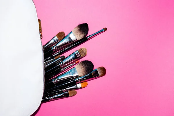 Make up brushes spilling out of a white cosmetics bag on pink background.Top view. Flat lay. Beauty concept