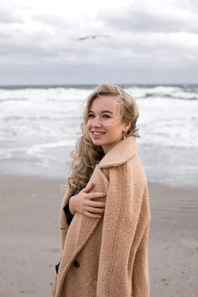 秋天的照片拍摄与一个美丽的女孩在海边 穿着外套的金发姑娘 多云的天气 背景中的波浪 — 图库照片