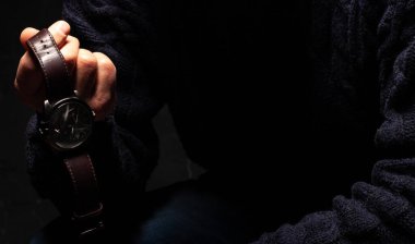 Bir adam bir saati sıkıca elinde tutar. Saatin kaç olduğunu gösteriyor. Siyah arka plan, süveter giyen yetişkin bir adam..
