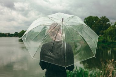 Arkadan şemsiyesi olan bir kız. Şemsiye şeffaf, yağmurlu hava. Güzel doğa, bir göl ve bir sürü ağaç..