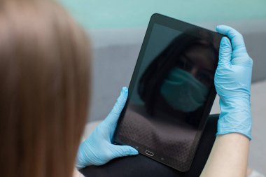 Eğer tablet daha fazla insan tarafından kullanılırsa, güvenlik için lastik koruyucu eldivenler takmalısınız ki enfeksiyon kapmasın. Bir lise mezunu elinde tabletle yatağında oturuyor. Plastik eldivenler ve koruyucu bir maskeyle..