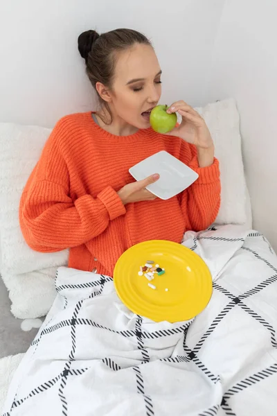 一个少年穿着一件橙色毛衣坐在被子下面的床上 他在吃一个青苹果 — 图库照片