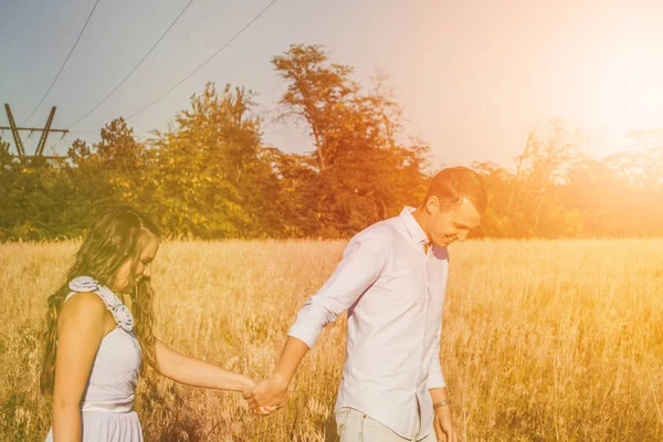 Νεαρό ζευγάρι ερωτευμένο να κρατιέται χέρι χέρι και να χαμογελάει στο χωράφι. Ο άντρας οδηγεί τη γυναίκα μπροστά. Εξωτερικό πορτρέτο του όμορφου ρομαντικού ζευγαριού με ελαφρά ρούχα που ποζάρουν το καλοκαίρι. Φαινόμενο φωτοβολίδας — Φωτογραφία Αρχείου