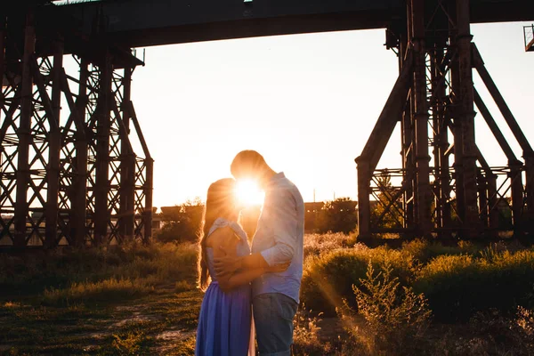 Young couple in love hugs on sunset amidst a bridge under construction. Man hugs woman closely. Outdoor portrait of beautiful romantic couple in light clothes in sun rays. Copy space — Stock Photo, Image