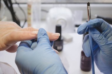 Mavi lastik eldivenli manikür güzellik salonu ustası, diğer enstrümanların beyaz arka planına siyah fırçalı, şeffaf jel katmanı uygular. Sağlıklı ve özenli. Manikür için profesyonel araç