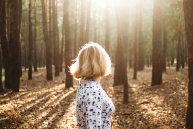 Sarışın, kısa saçlı, beyaz elbiseli, doğa kökenli genç bir kadın. Dişi, çam ormanında kameraya karşı duruyor. Kız ağaçlardan, havadan ve manzaradan hoşlanıyor. Açık hava portresi. Moda yaz fotoğrafı.