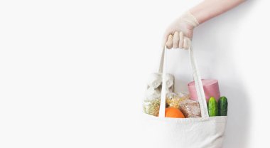 Lastik eldivenler, içinde yumurta, karabuğday, yulaf, bezelye, salatalık, portakal, makarna ve tuvalet kağıdı olan bez torbaları. Beyaz arka planda yemek teslimatı. Koronavirüs karantinası için bağış. Boşluğu kopyala