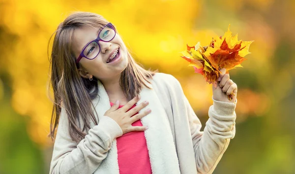 Herfst. Portret van een lachende jong meisje die in haar hand een boeket van herfst esdoorn houdt bladeren. Pre-teen jong meisje met bril en tanden bretels — Stockfoto