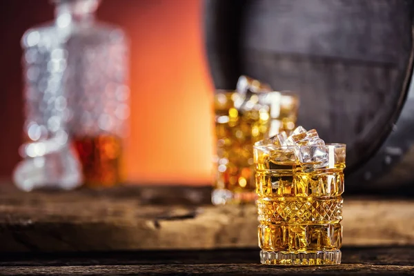 Whiskey. Twee bekers vol drank whiskey brandewijn of cognac met ijsblokjes in retro stijl. Oude eiken vat op de achtergrond — Stockfoto