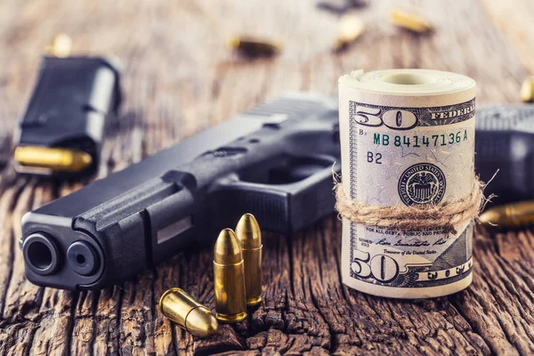 Gun and money. 9 mm pistol gun bullets strewn and roll dollar banknotes on rustic oak table — Stock Photo, Image