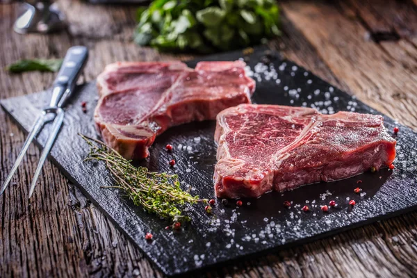 Carne de res T-bone. Carne cruda de res T-bone en pizarra con sal pimienta hierbas carnicero y tenedor — Foto de Stock