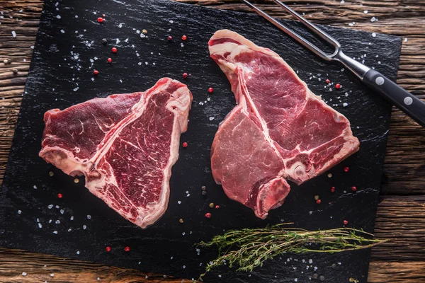 Carne de res T-bone. Carne cruda de res T-bone en pizarra con sal pimienta hierbas carnicero y tenedor — Foto de Stock