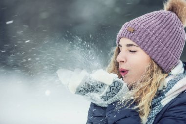 Güzel, genç bir kadın, kalın giysiler içinde kar üflüyor.