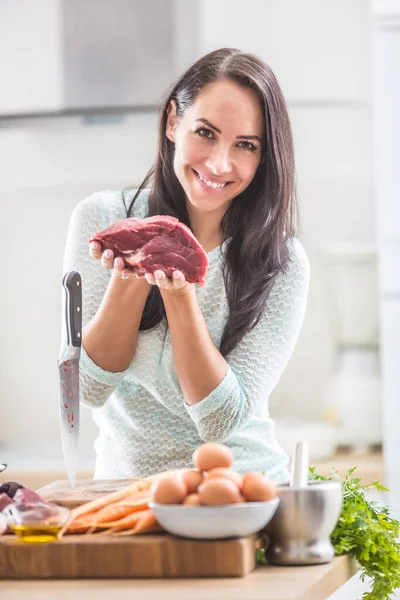 Cheerful female cook in kitchen holding raw beef meat