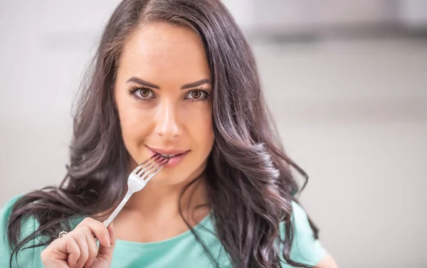 A beautiful young woman with a fork in her mouth is thinking with the temptation to eat.