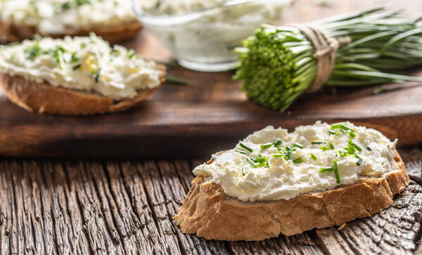 Slices of crusty bread with a cream cheese spread and freshly cut chives on a vintage wooden cutting board.