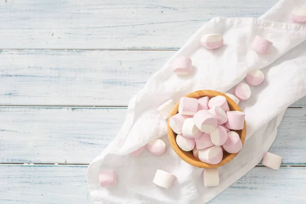 Tampilan atas mangkuk kayu penuh marshmallow merah muda dan putih dengan beberapa tersebar di sekitar pada kain meja putih dan permukaan kayu putih — Stok Foto