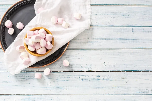 Ovanifrån av en träskål full av rosa och vit marshmallows med några utspridda runt på en vit bordsduk, svart bricka och vit träyta — Stockfoto