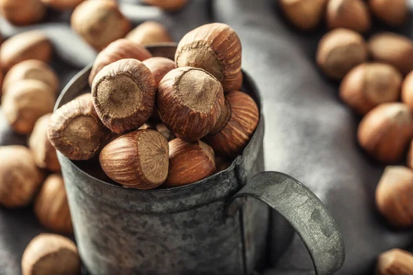 Närbild av en vintage metallic mugg full av hasselnötter med mer nötter på en textilduk runt den — Stockfoto