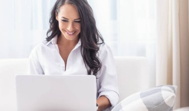 Beyaz gömlekli çekici bir kadın oturma odasında defteriyle çalışıyor, gülümsüyor..