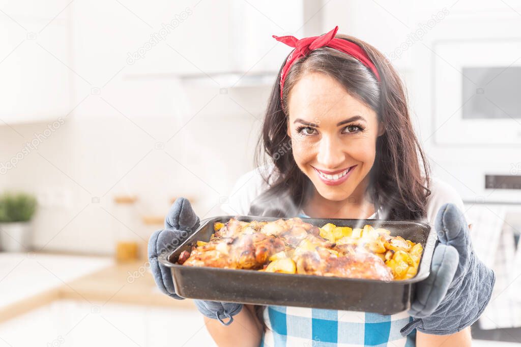 Beautiful wife holding a steaming hot tray in the kitchen, holding baked chicken and potatoes in her oven gloves, wearing apron and a red ribbon in her hair, smiling and looking into the camera.