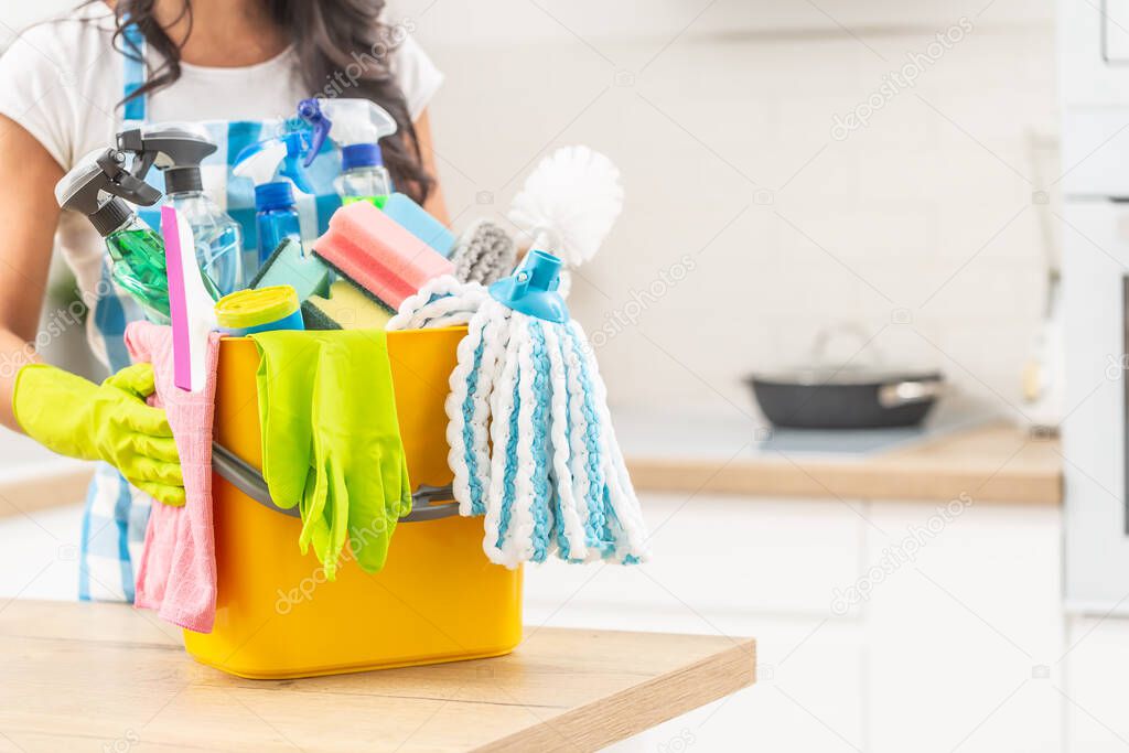 Bucket full of house clenaing stuff on a kitchen desk with a female holding it in rubber gloves. 