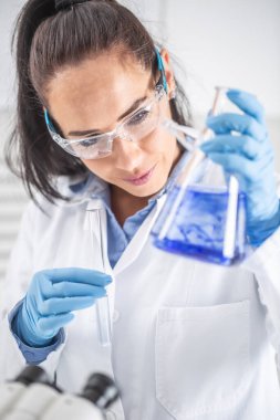 Koruyucu gözlük takan kadın kimyager deney tüpü tutarken gülümseyip konik şişenin içine mavi sıvıyı karıştırıyor. Coronavirus COVID-19 konsepti.