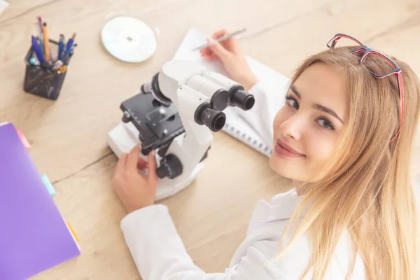 Jovem Loira Olhando Para Câmera Enquanto Trabalhava Com Microscópio Escrevendo — Fotografia de Stock