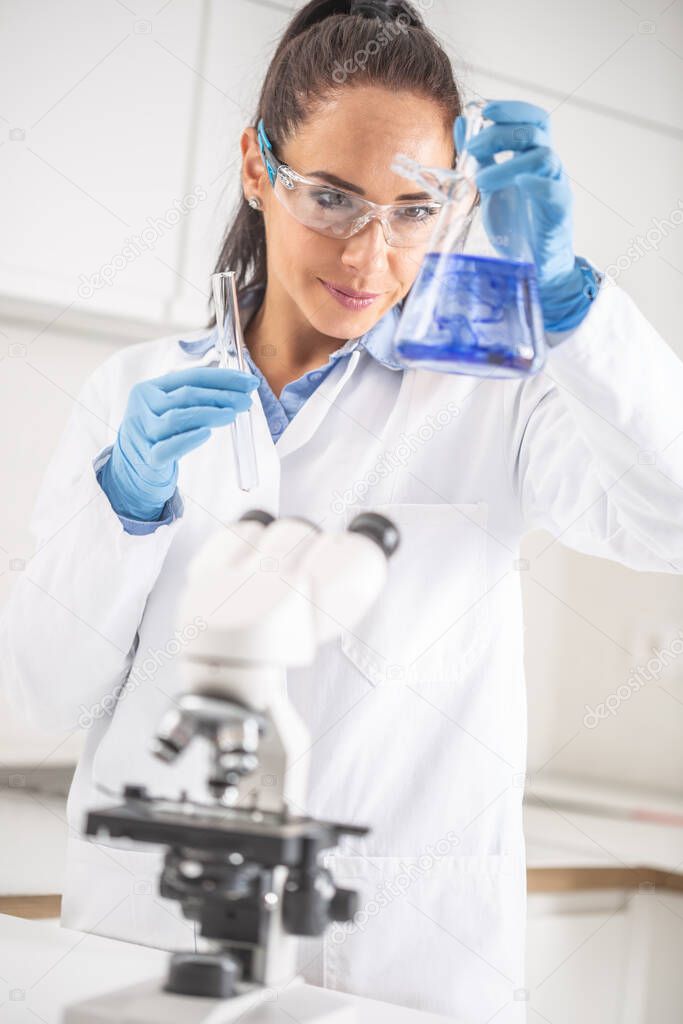Female chemist wearing protective goggles smiles when holding a test-tube, mixing blue liquid inside the conical flask. Coronavirus COVID-19 concept.