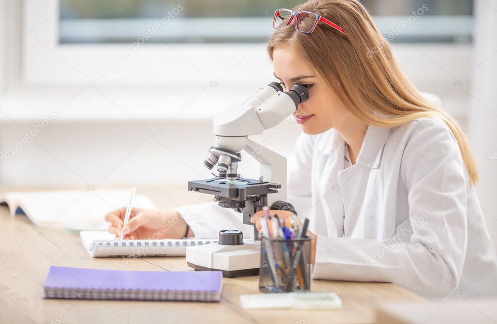 Beautiful young student girl researcher looking into a microscope, writing down notes.