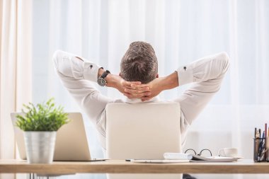 İşadamı, elleri başının arkasında, ofis koltuğunda oturur, sırtını döner ve dinlenir..
