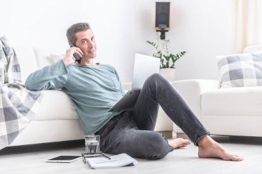 Günlük kıyafet giyen bir adam apartmanın zemininde oturuyor, önünde bir not defteriyle cep telefonundan arıyor..