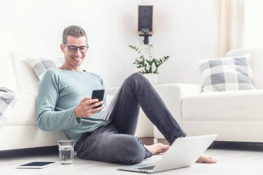 Evde çalışırken cep telefonunda gülümseyen adam mesajları, oturma odasında yerde oturuyor..