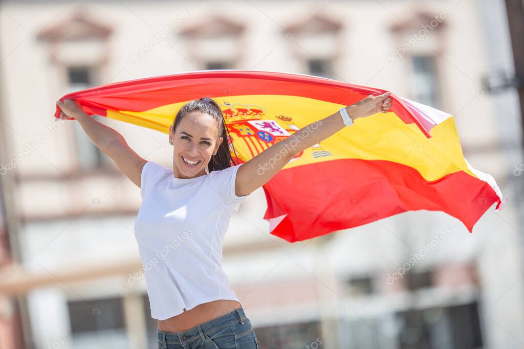 Beautiful girl holds a Spanish flag in her hands behind her, cheering on a street.