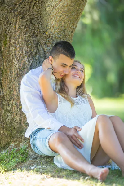 年轻的恋人坐在树下 女孩抚摸着男孩的脖子 在炎炎夏日微笑着 — 图库照片