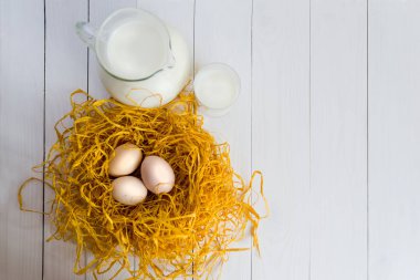 Üç tavuk yumurtası ile bir sürahi ve bardak süt beyaz ahşap zemin üzerinde dekoratif yuvada. Doğal beslenme, tarım, Tavukçuluk kavramı