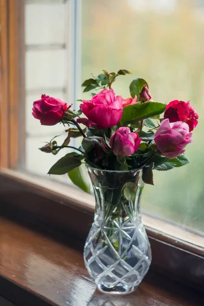 Hausgemachte Rosen. Strauß in einer Kristallvase an einem Holzfenster. das Konzept des ruhigen und gemütlichen Dorflebens, der Gartenarbeit und der Umweltfreundlichkeit lizenzfreie Stockfotos