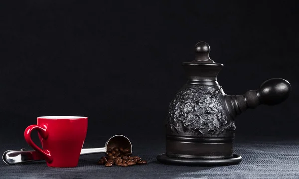 A coffeepot from black clay, red coffee cup and steel measuring spoon with roasted coffee beans on a background of rough dark blue fabric.