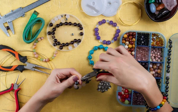 Jewelry making. Making a bracelet of colorful beads. Female hands with a tool on a yellow background.